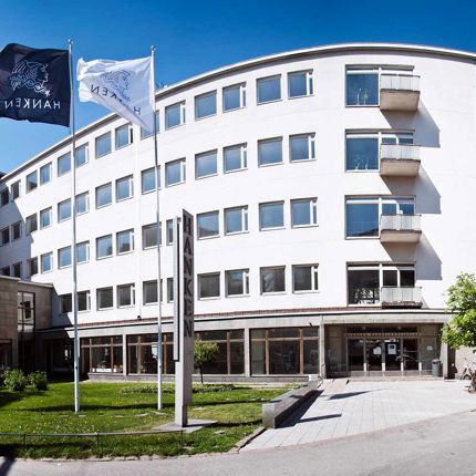 The facade of Hanken School of Economics in Helsinki, a white building with large windows, flagpoles, and a green outdoor area.