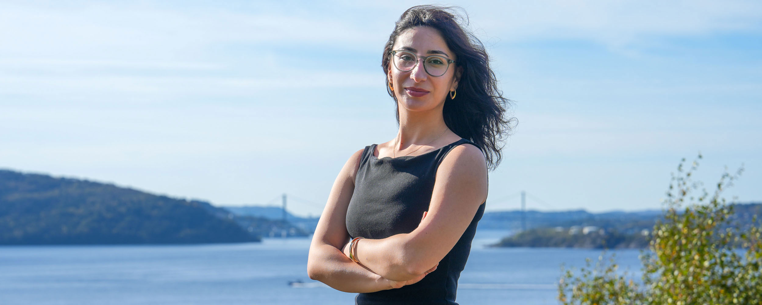 Portrett av en smilende kvinne med briller, som står utendørs med havet i bakgrunnen på NHH sitt campus