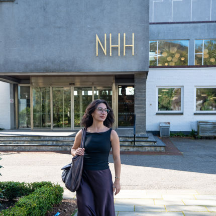 En kvinne med briller, svart topp, mørkt skjørt og en skulderveske går på campuset til Norges Handelshøyskole (NHH), med NHH-skiltet synlig på bygningen bak henne
