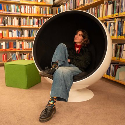 A woman sits in a white spherical chair in a library, surrounded by bookshelves.
