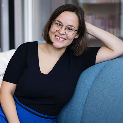 A photo of a woman with dark medium long hair sitting on a couch smiling at the camera. 