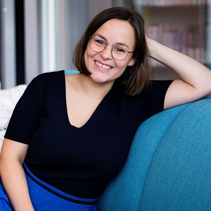 Portrettbilde av en dame med mørkt halvlangt hår og briller som sitter i en turkis sofa og smiler til kamera.